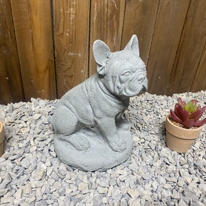 May include: A gray concrete statue of a French bulldog sitting on a bed of gray gravel.