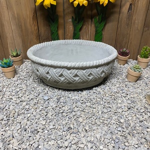 May include: A gray concrete planter with a woven design. The planter is sitting on a bed of gray gravel. Three yellow metal sunflowers are mounted on a wooden wall behind the planter.