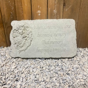 May include: A gray stone garden memorial with a carved angel and the text "When someone you love becomes a memory That memory becomes a treasure..."