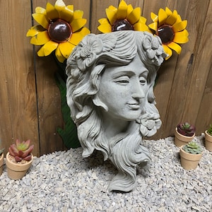 May include: A gray concrete planter shaped like a woman's head with long hair. The planter has a flower design around the head and a flower on the side. Three yellow metal sunflowers are above the planter.