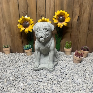 May include: A gray concrete pug dog statue sitting on a bed of gray gravel with three yellow sunflowers behind it.