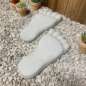 Op de afbeelding: Twee grijze betonnen voetafdrukken op een bed van kleine witte grindstenen. Op de achtergrond staan ​​drie kleine potplanten met groene en rode bladeren.