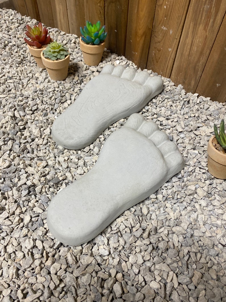 Op de afbeelding: Twee grijze betonnen voetafdrukken op een bed van kleine witte grindstenen. Op de achtergrond staan ​​drie kleine potplanten met groene en rode bladeren.