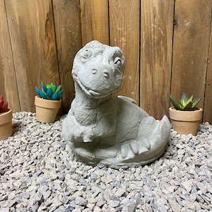 May include: A small, gray concrete dinosaur statue with a friendly expression. The dinosaur is sitting on a bed of gray gravel.