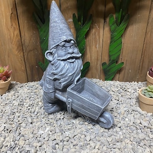 May include: A gray stone garden gnome figurine with a white beard and a wide smile, pushing a gray wheelbarrow. The gnome is wearing a pointed hat and is standing on a bed of gray gravel.