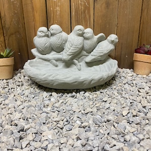May include: A gray concrete bird bath with four birds perched on the rim. The birds are facing each other and have detailed features. The bird bath is sitting on a bed of gray gravel.