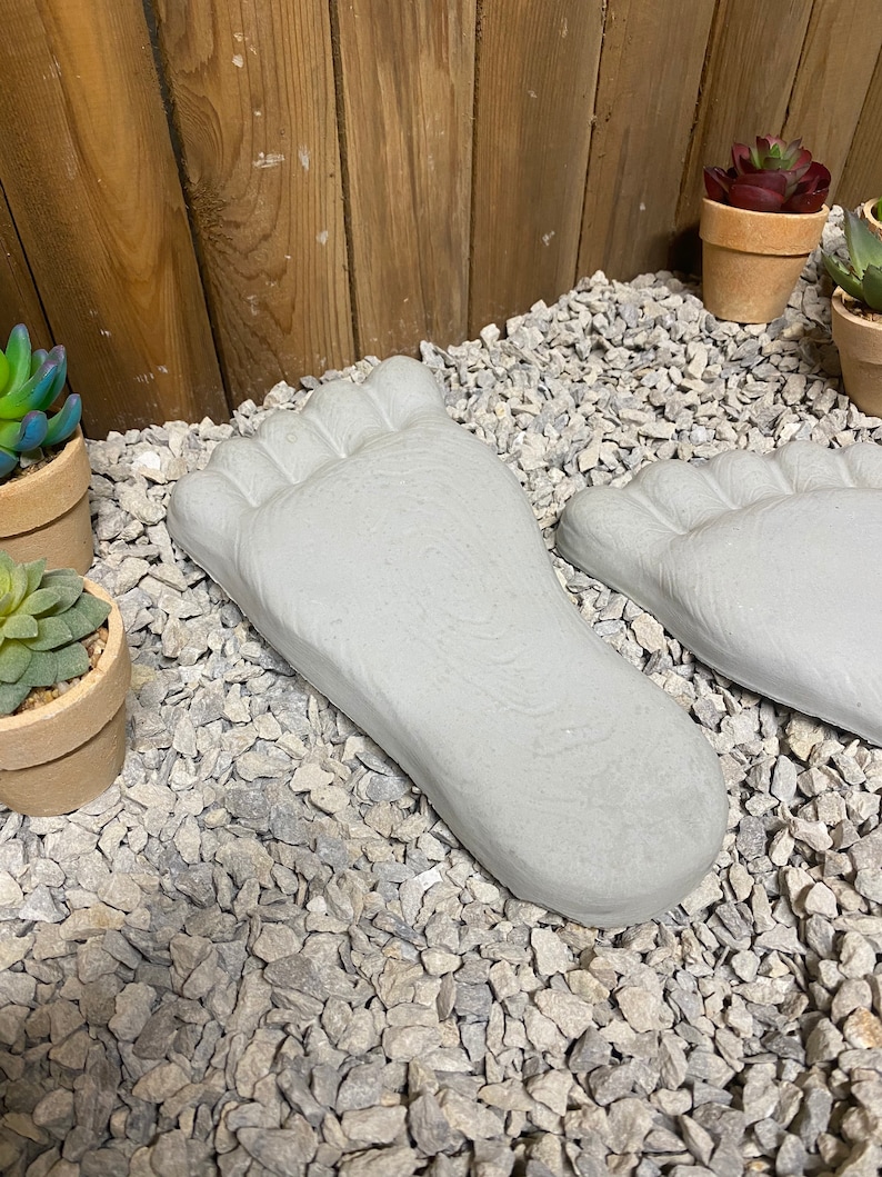 Op de afbeelding: Twee grijze betonnen voetvormige stapstenen op een bed van grijs grind. De stenen zijn getextureerd om op een blote voet te lijken.