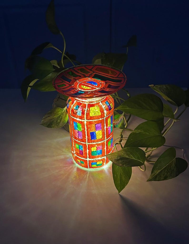 May include: A decorative, illuminated jar with a mosaic design. The jar is primarily orange with colorful squares and a matching lid. The light inside casts a colorful glow. Green leaves are in the background.