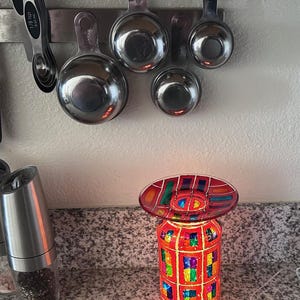 May include: A colorful, stained-glass style electric candle warmer with a red and multi-colored design. The warmer has a cylindrical base and an oval top. Stainless steel measuring cups are mounted on a magnetic strip in the background.