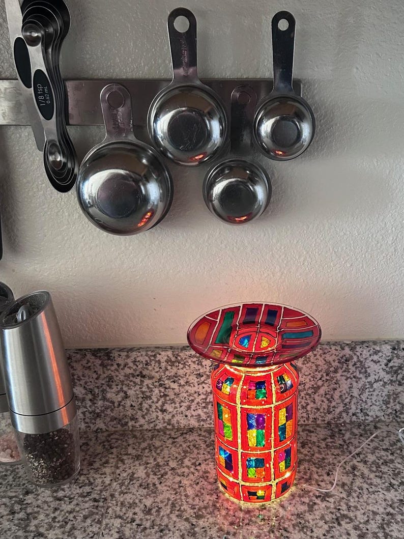 May include: A colorful, stained-glass style electric candle warmer with a red and multi-colored design. The warmer has a cylindrical base and an oval top. Stainless steel measuring cups are mounted on a magnetic strip in the background.