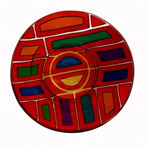 May include: A round, red decorative plate with a colorful geometric design. The plate features a central gold circle with a blue and gold inner design, surrounded by red, orange, green, blue, and purple shapes.