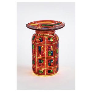 May include: A decorative, stained-glass style candle holder. The red glass jar is adorned with a grid pattern of colorful squares and a wide, circular top. The candle holder is illuminated from within, creating a warm glow.