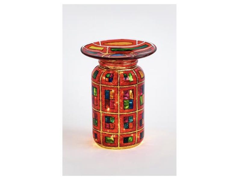 May include: A decorative, stained-glass style candle holder. The red glass jar is adorned with a grid pattern of colorful squares and a wide, circular top. The candle holder is illuminated from within, creating a warm glow.