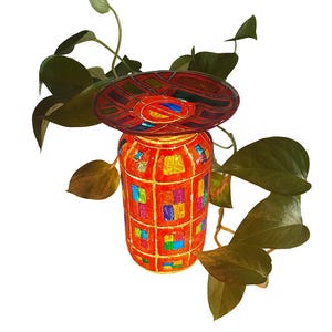May include: A decorative, handmade jar with a matching lid, featuring a vibrant mosaic design in red, orange, blue, and yellow. The jar is adorned with colorful squares and a geometric pattern. Green leaves from a plant surround the jar.