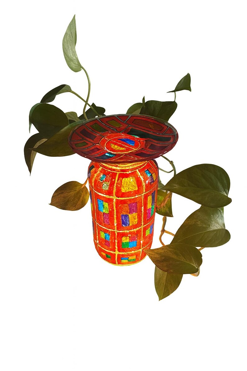 May include: A decorative, handmade jar with a matching lid, featuring a vibrant mosaic design in red, orange, blue, and yellow. The jar is adorned with colorful squares and a geometric pattern. Green leaves from a plant surround the jar.