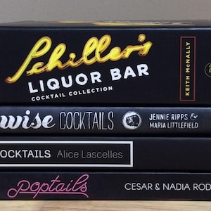 May include: A stack of four black cocktail recipe books. The top book is titled "Schiller's Liquor Bar Cocktail Collection" in gold lettering. Other titles include "wise COCKTAILS", "TEN COCKTAILS", and "Poptails".