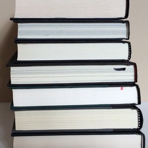May include: A stack of seven hardcover books with black spines and white pages. The books are arranged vertically, with the spines facing outward. The edges of the pages are white, and the covers are black.