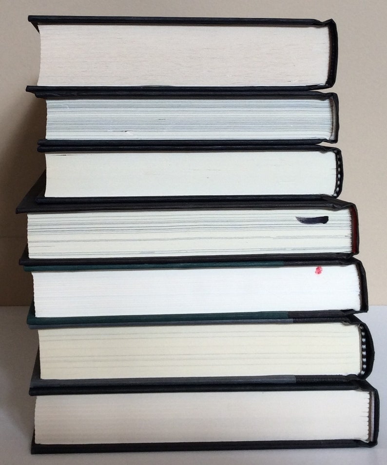 May include: A stack of seven hardcover books with black spines and white pages. The books are arranged vertically, with the spines facing outward. The edges of the pages are white, and the covers are black.