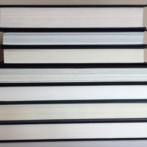 May include: A stack of hardcover books with black spines and white pages. The books are arranged in a neat pile, with the pages facing upwards. The edges of the pages are a light cream color. The books are of varying thicknesses.