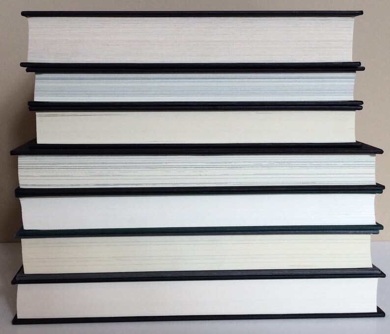 May include: A stack of hardcover books with black spines and white pages. The books are arranged in a neat pile, with the pages facing upwards. The edges of the pages are a light cream color. The books are of varying thicknesses.