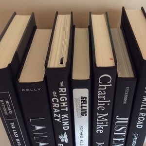 May include: A collection of hardcover books with black covers and cream-colored pages. Titles and author names are visible on the spines, including "The Last Magazine" and "Grist Mill Road". The books are arranged in a row, with varying thicknesses.