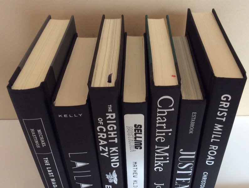May include: A collection of hardcover books with black covers and cream-colored pages. Titles and author names are visible on the spines, including "The Last Magazine" and "Grist Mill Road". The books are arranged in a row, with varying thicknesses.