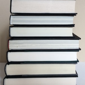 May include: A stack of seven hardcover books with black covers and white pages. The books are neatly arranged, with the spines facing the viewer. The edges of the pages are visible, and the books appear to be in good condition.