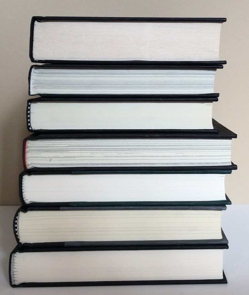 May include: A stack of seven hardcover books with black covers and white pages. The books are neatly arranged, with the spines facing the viewer. The edges of the pages are visible, and the books appear to be in good condition.