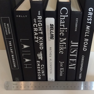 May include: A collection of hardcover books with black covers and white lettering. Titles include "The Last Magazine," "Lailah," "The Right Kind of Crazy," "Selling Nostalgia," "Charlie Mike," "Just Eat," and "Grist Mill Road." A ruler is at the bottom.