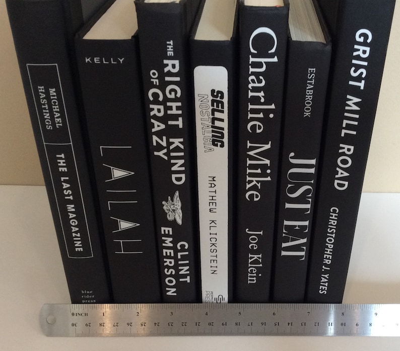 May include: A collection of hardcover books with black covers and white lettering. Titles include "The Last Magazine," "Lailah," "The Right Kind of Crazy," "Selling Nostalgia," "Charlie Mike," "Just Eat," and "Grist Mill Road." A ruler is at the bottom.