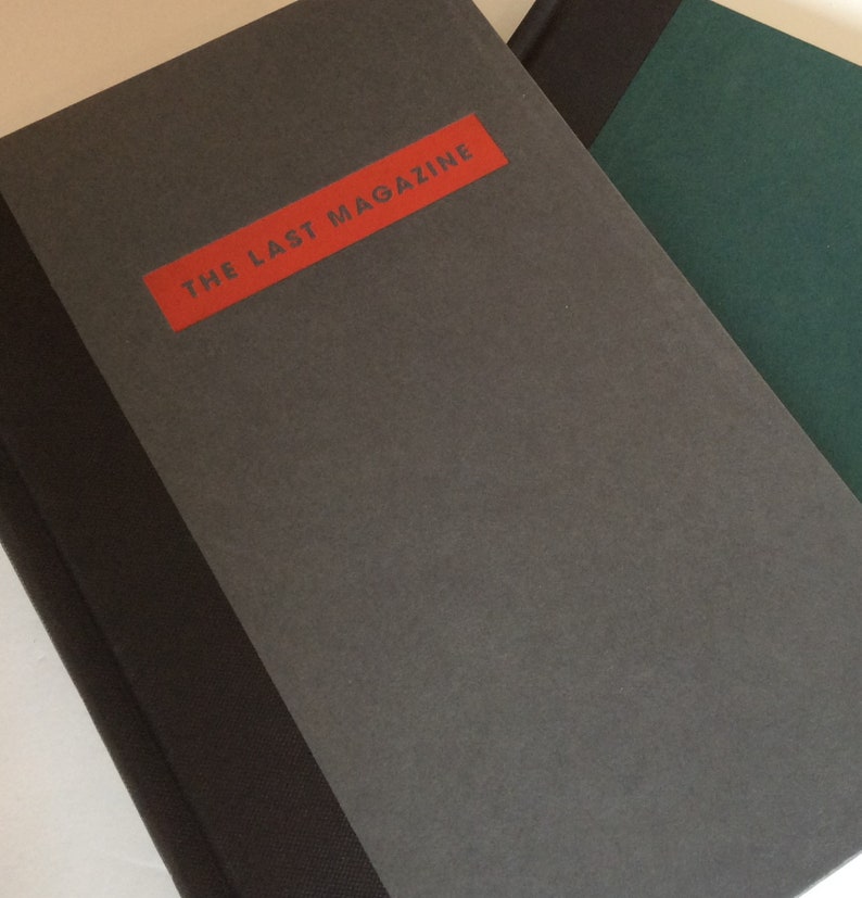 May include: A stack of books with a gray cover and a red rectangular label that reads "THE LAST MAGAZINE". The spine is black. Another book with a green cover is partially visible.