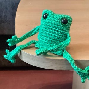 May include: A green crocheted frog with black eyes sitting on a wooden surface.