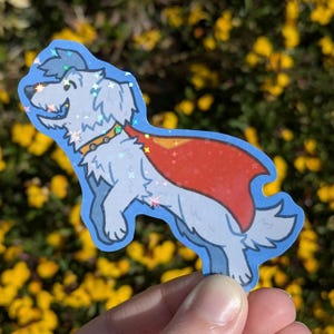 May include: A sticker of a cartoon dog wearing a red cape and yellow collar. The dog is light grey with a blue outline and a glittery, holographic finish. The background is blurred with yellow flowers.