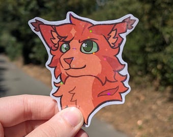 May include: A red and orange cartoon cat head sticker with green eyes. The sticker has a holographic finish.