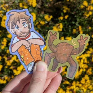 Puede incluir: Dos pegatinas holográficas. Una presenta a un hombre de dibujos animados con gafas, que lleva una camiseta blanca con ribetes rojos y pantalones naranjas. La otra pegatina es una criatura marrón con forma de roca con los brazos levantados en señal de paz.