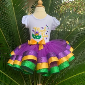 Op de afbeelding: Een witte top met korte mouwen, met de tekst "Mardi Gras" en een maskerontwerp, gecombineerd met een paarse tutu-rok. De rok heeft lagen paarse, gouden en groene linten. Een gouden strik siert de taille. De outfit staat op een mannequin.