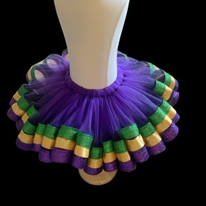 Op de afbeelding: Een paarse tutu met lagen tule en decoratieve linten. De linten hebben afwisselend de kleuren groen, goud en paars. De tutu wordt getoond op een witte mannequin.