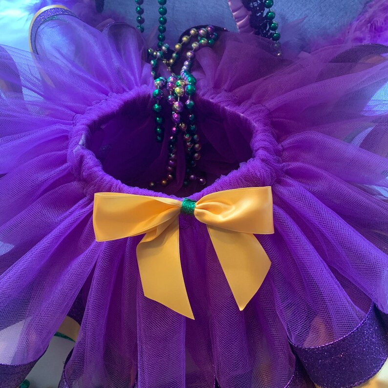 Op de afbeelding: Een paarse tutu met een gele strik en groene accenten. De tutu is gemaakt van lagen tule en heeft een glitterrand. Een streng Mardi Gras-kralen is over de tutu gedrapeerd.