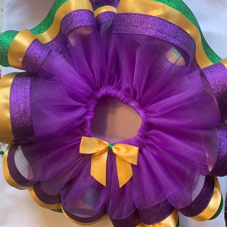 Op de afbeelding: Een paarse tutu met gouden en groene accenten. De tutu heeft lagen paarse tule, gouden satijnen lint en groen lint. Een gouden strik met een groen centrum siert de voorkant. De randen zijn afgewerkt met paarse glitter.