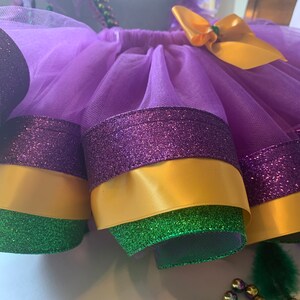 Op de afbeelding: Een paarse tule tutu rok met lagen glinsterende paarse, gouden en groene afwerking. Een gouden strik is aan de taille bevestigd. De rok is ontworpen met Mardi Gras kleuren.