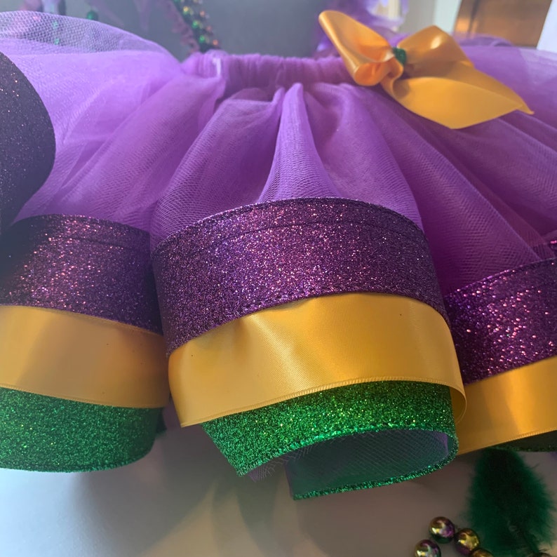 Op de afbeelding: Een paarse tule tutu rok met lagen glinsterende paarse, gouden en groene afwerking. Een gouden strik is aan de taille bevestigd. De rok is ontworpen met Mardi Gras kleuren.
