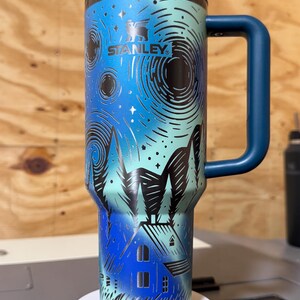 Puede incluir: Un termo de viaje Stanley con asa y tapa azules. El termo presenta un dise&ntilde;o de cielo nocturno con una casa y &aacute;rboles. El logotipo de Stanley es visible en la parte delantera. El termo es cil&iacute;ndrico y mide unos 23 cm de alto.