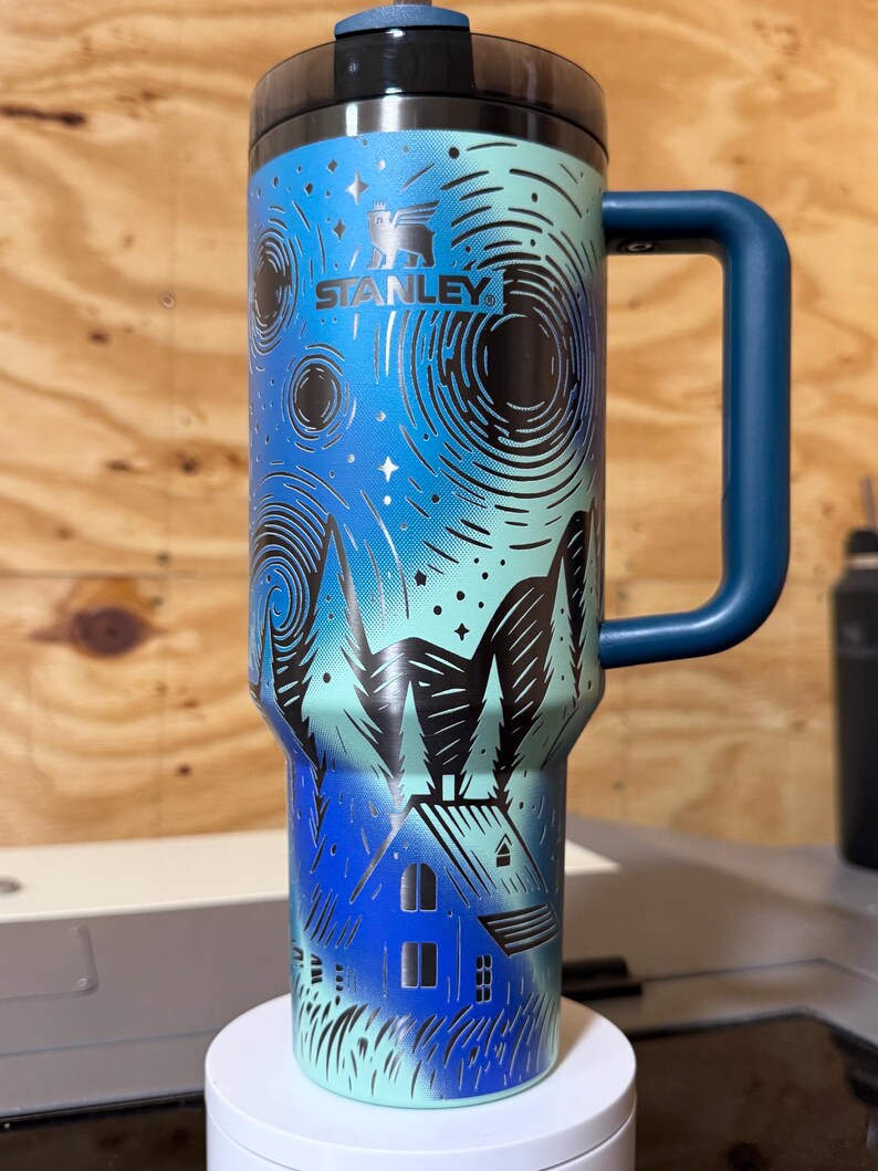 Puede incluir: Un termo de viaje Stanley con asa y tapa azules. El termo presenta un dise&ntilde;o de cielo nocturno con una casa y &aacute;rboles. El logotipo de Stanley es visible en la parte delantera. El termo es cil&iacute;ndrico y mide unos 23 cm de alto.