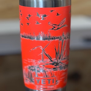 May include: A stainless steel tumbler with a bright orange finish. The tumbler features a detailed black illustration of ducks in flight over a marsh scene. The word "YETI" is visible at the bottom of the tumbler.