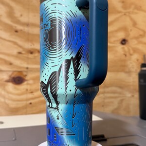 Puede incluir: Un vaso Stanley de color verde azulado y azul con asa. El vaso presenta un dise&ntilde;o de monta&ntilde;a y casa con la palabra "Stanley" visible. La tapa y el asa son de color azul oscuro. Una pajita est&aacute; insertada en la tapa.