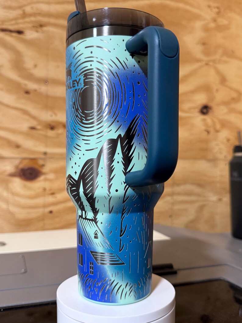 Puede incluir: Un vaso Stanley de color verde azulado y azul con asa. El vaso presenta un dise&ntilde;o de monta&ntilde;a y casa con la palabra "Stanley" visible. La tapa y el asa son de color azul oscuro. Una pajita est&aacute; insertada en la tapa.