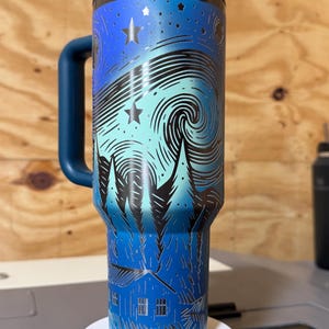 Puede incluir: Un termo de viaje de acero inoxidable con un dise&ntilde;o de noche estrellada azul y turquesa. El termo presenta un asa y tapa azul oscuro, y una ilustraci&oacute;n detallada de un cielo nocturno arremolinado con estrellas y &aacute;rboles.
