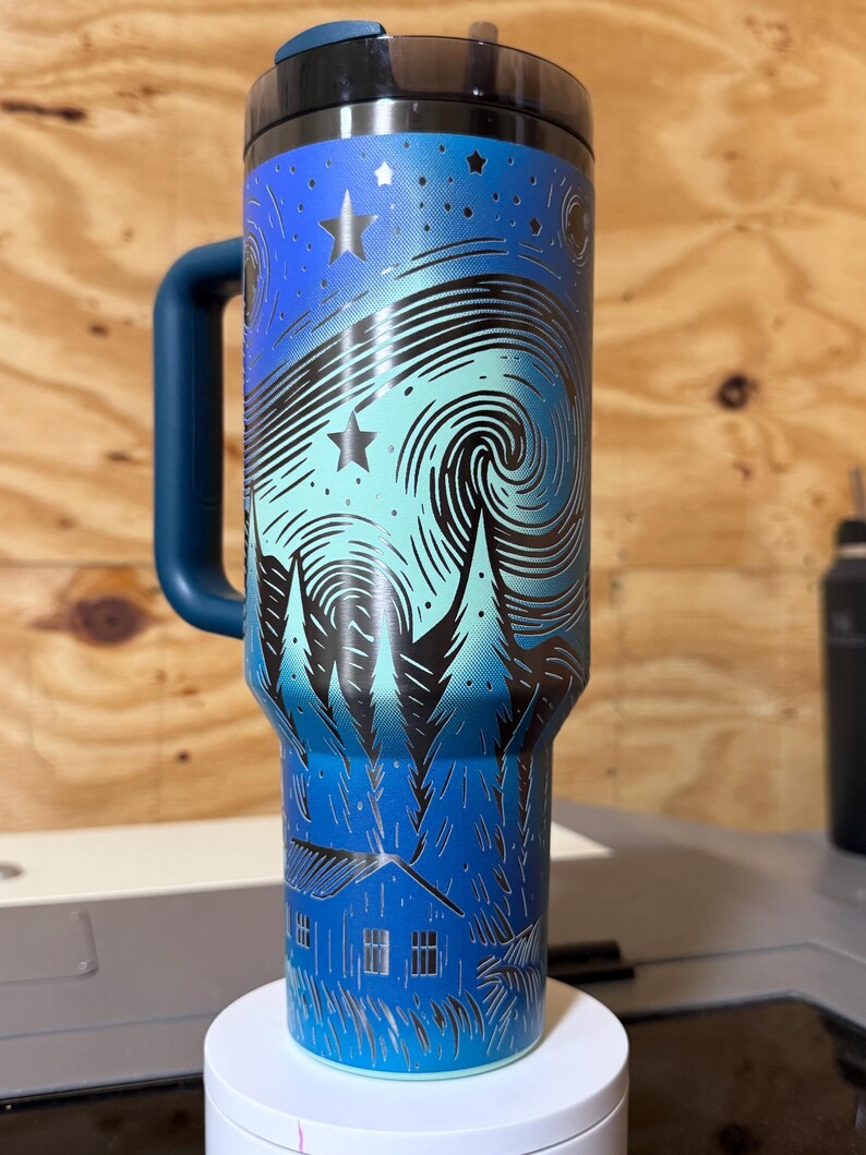 Puede incluir: Un termo de viaje de acero inoxidable con un dise&ntilde;o de noche estrellada azul y turquesa. El termo presenta un asa y tapa azul oscuro, y una ilustraci&oacute;n detallada de un cielo nocturno arremolinado con estrellas y &aacute;rboles.