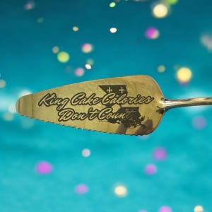 May include: Gold-colored cake server with a serrated edge. The server has the text "King Cake Calories Don't Count" engraved on it. The server is against a blue background with out-of-focus white and pink circles.