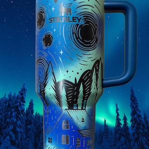 Puede incluir: Un termo de viaje Stanley azul y verde azulado con asa, con un dise&ntilde;o de paisaje invernal. El dise&ntilde;o incluye una casa, &aacute;rboles y patrones arremolinados, con el logotipo de Stanley. El termo tiene una tapa negra.